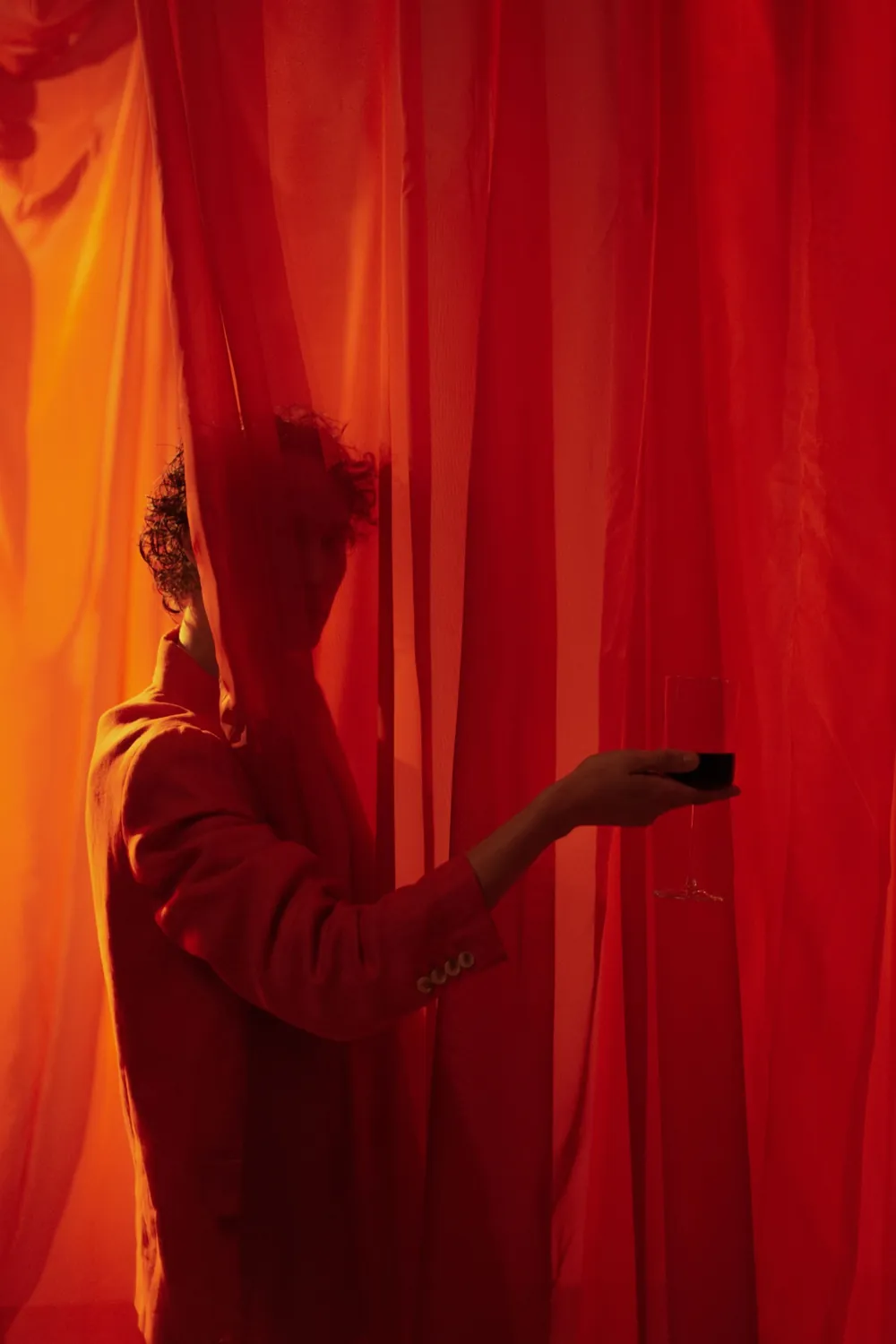 A figure half hidden behind red curtains, holding out a glass